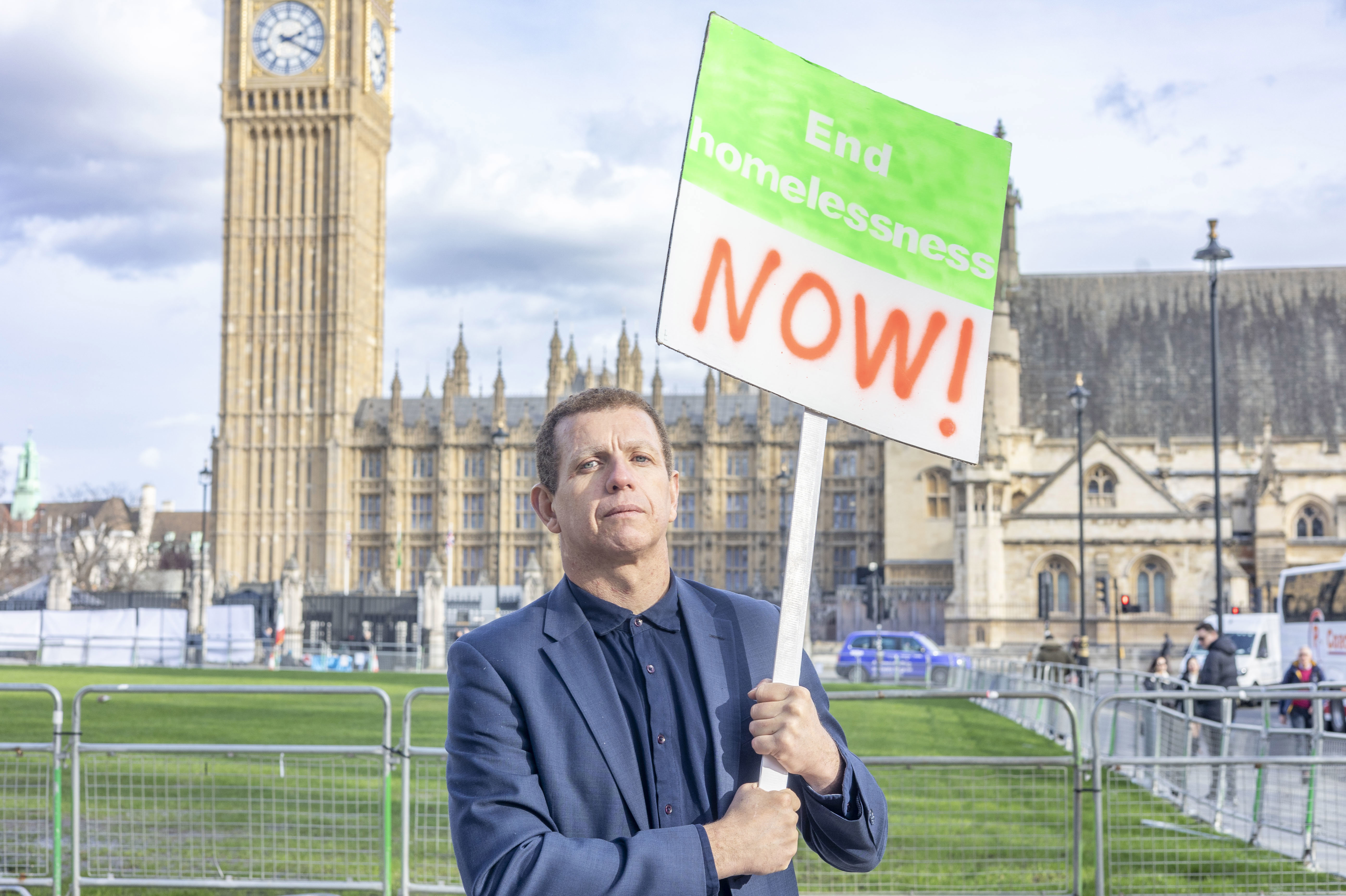 A man in his forties with light skin and wearing a blue suit stands in Parliament Square holding a sign that says 'End Homelessness Now!'