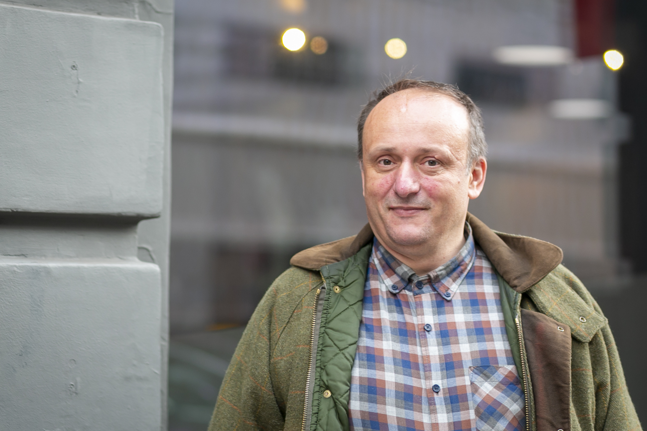 A white man with receding hair wearing a green coat and checked shirt stands in front of a window