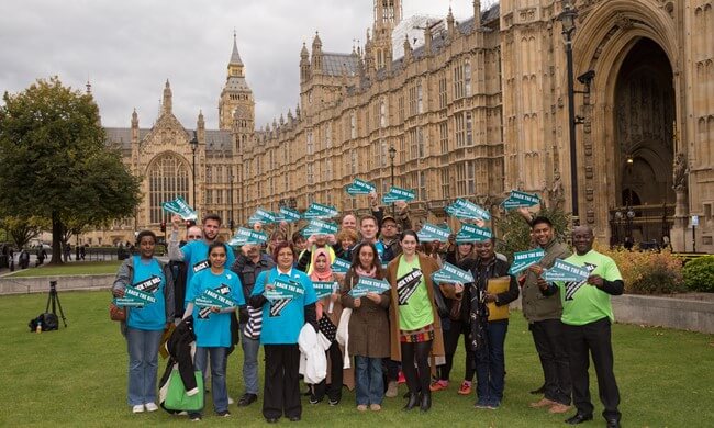 A group of Crisis campaigners stand outside the Houses of Parliament holding arrow shaped signs that say 'I back the bill'. 
