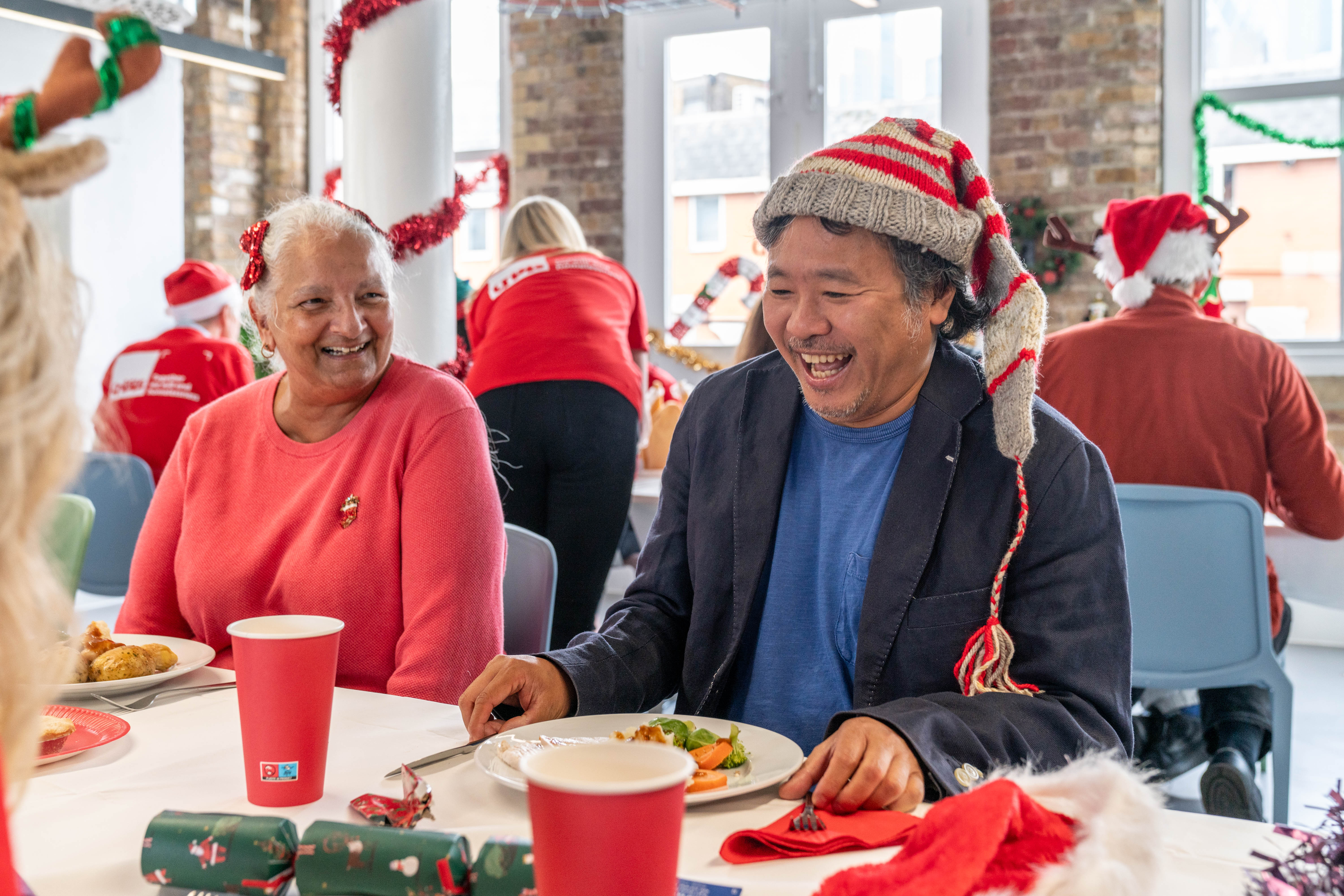 Two Crisis members are sat eating Christmas lunch together. They are smiling and laughing. The room has Christmas decorations up and the members are wearing festive hats.