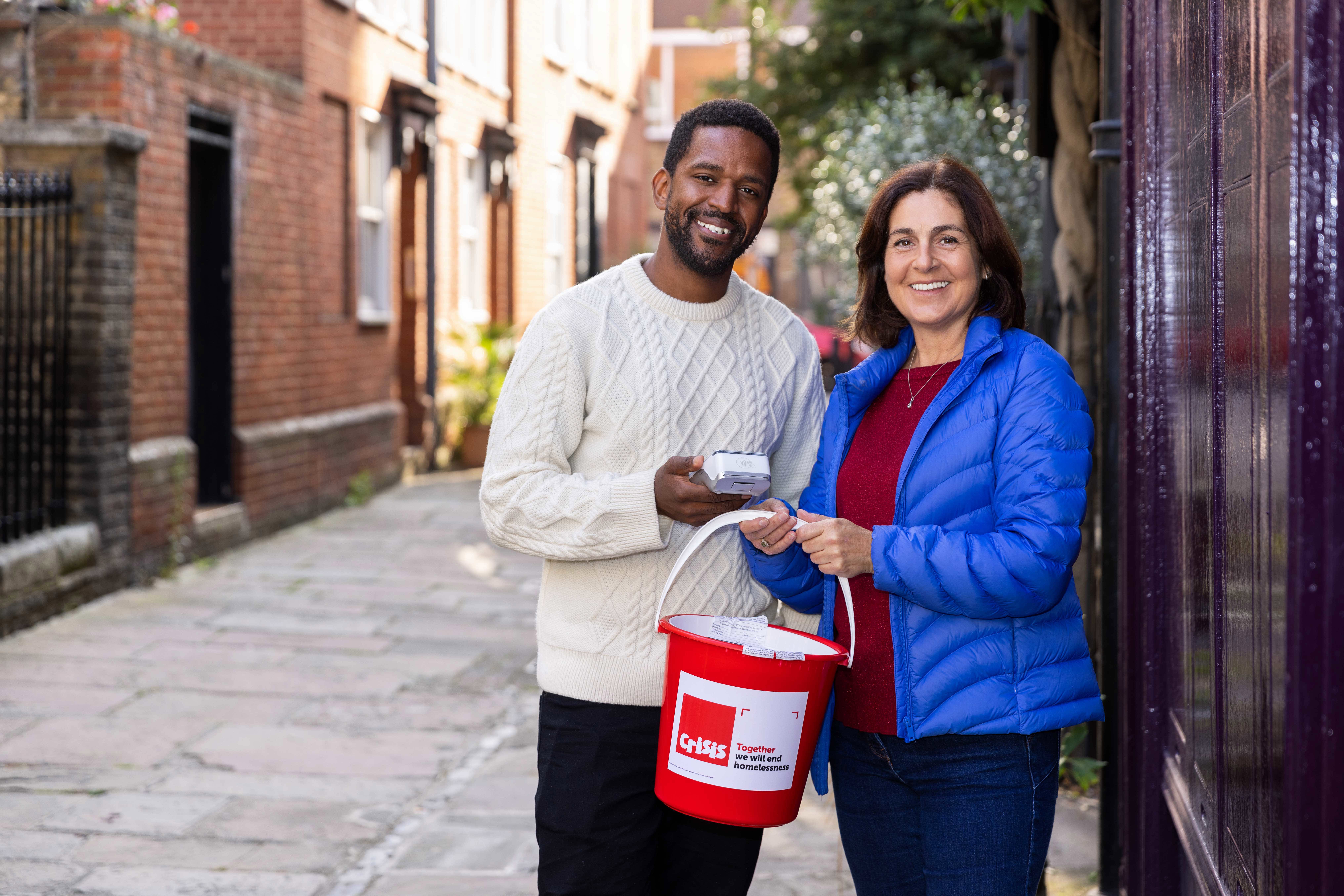 a man and woman standing in a street, holding a red Crisis collection bucket and smiling at the camera.