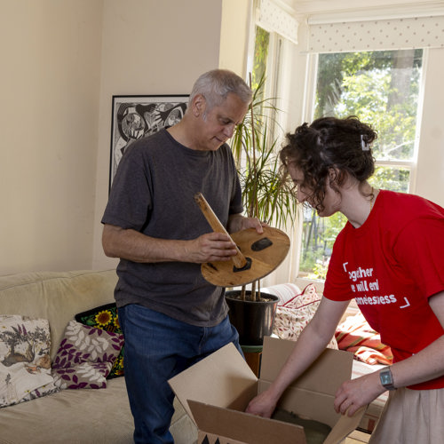 Massimo and a Crisis staff member are unpacking a box together in a bright living room. Through the window behind them, there is sunlit garden. 