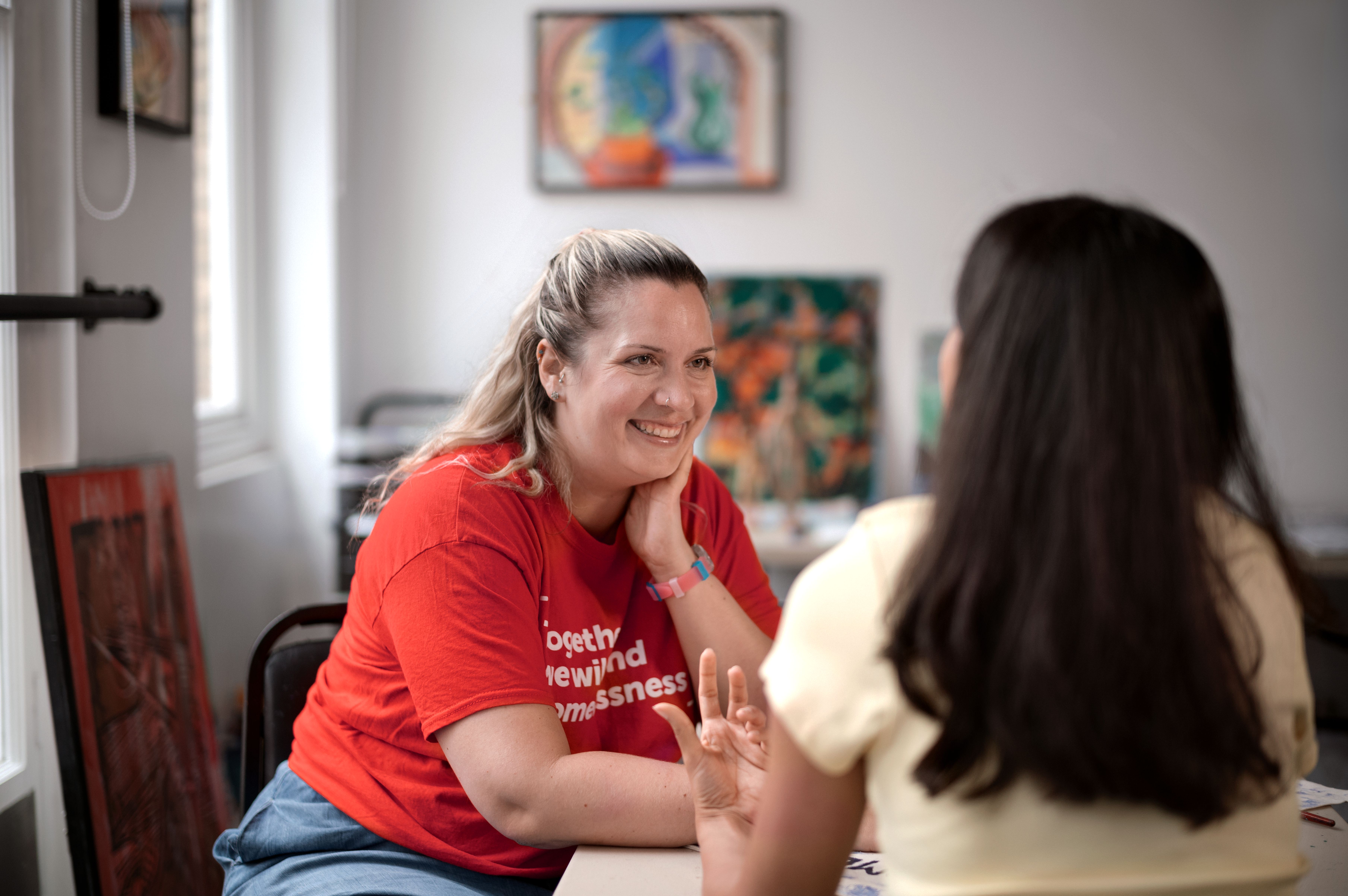 Anita is sat with her back to the camera. She is talking to a smiling member of Crisis staff. They are surrounded by art work in a brightly lit room.