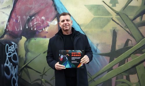Manoel stands in front of a wall with graffiti. He is holding his book 'Out in Parks'