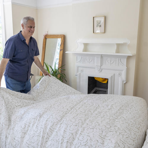 Massimo smiles while changing the covers on his bed. His bedroom is bright and tastefully decorated. 