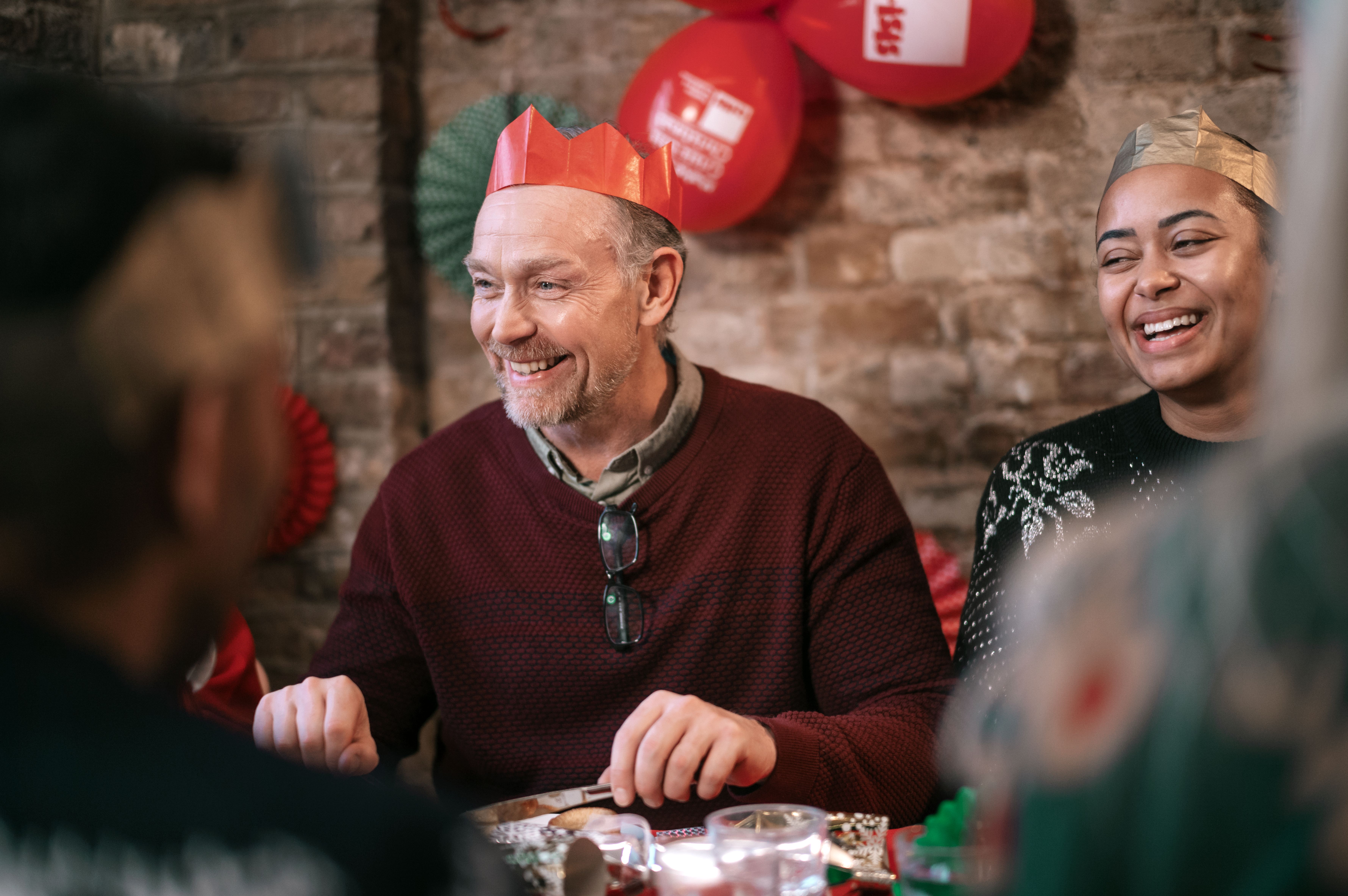 Simon is smiling broadly while wearing a paper crown from a Christmas cracker. Behind him there are Crisis branded balloons and Christmas decorations.
