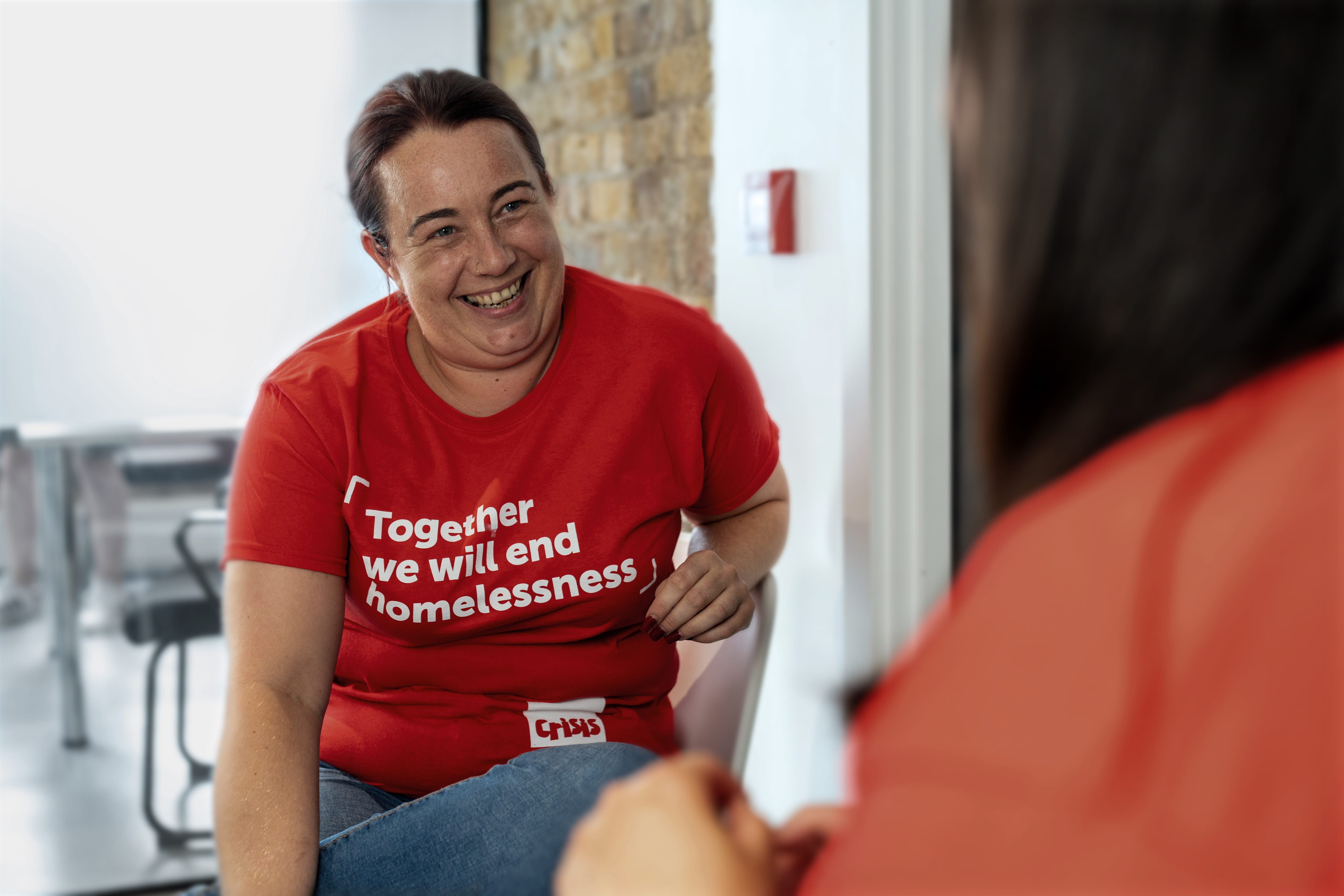 A woman wearing a red Crisis t-shirt talks to someone just out of shot. She is smiling and her body language is relaxed and friendly. 
