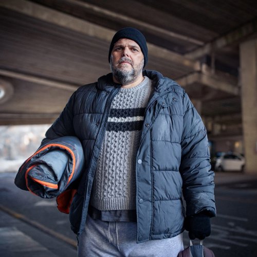 Gaz stands in a carpark in Winter. He is holding a rolled up sleeping bag and wearing a hat and gloves.