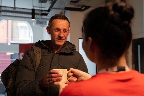 white man in warm clothing receiving a cup of tea from a crisis staff member