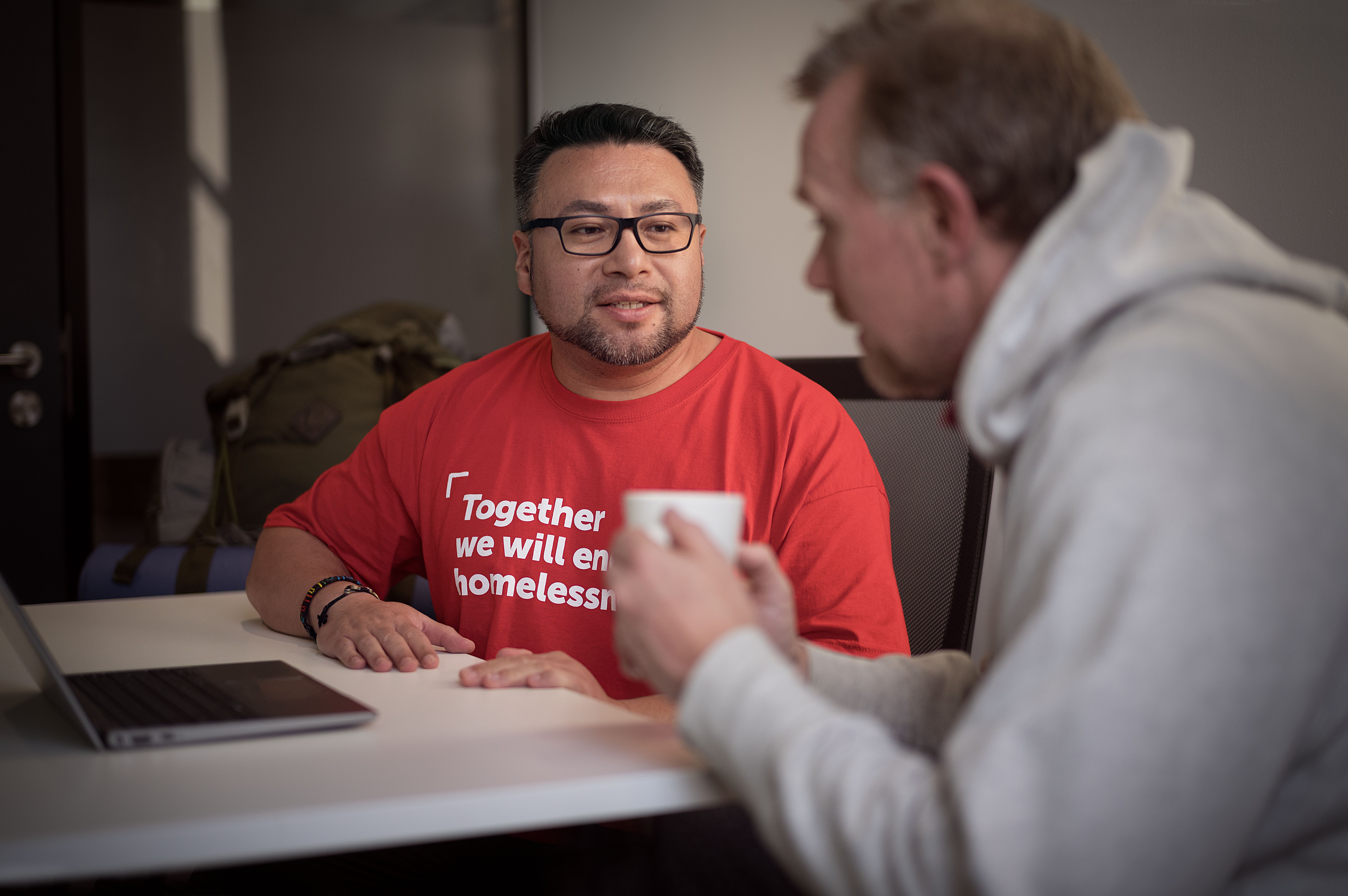 Simon is sat with a Crisis staff member. Simon is drinking a cup of tea and the staff member is listening to him.