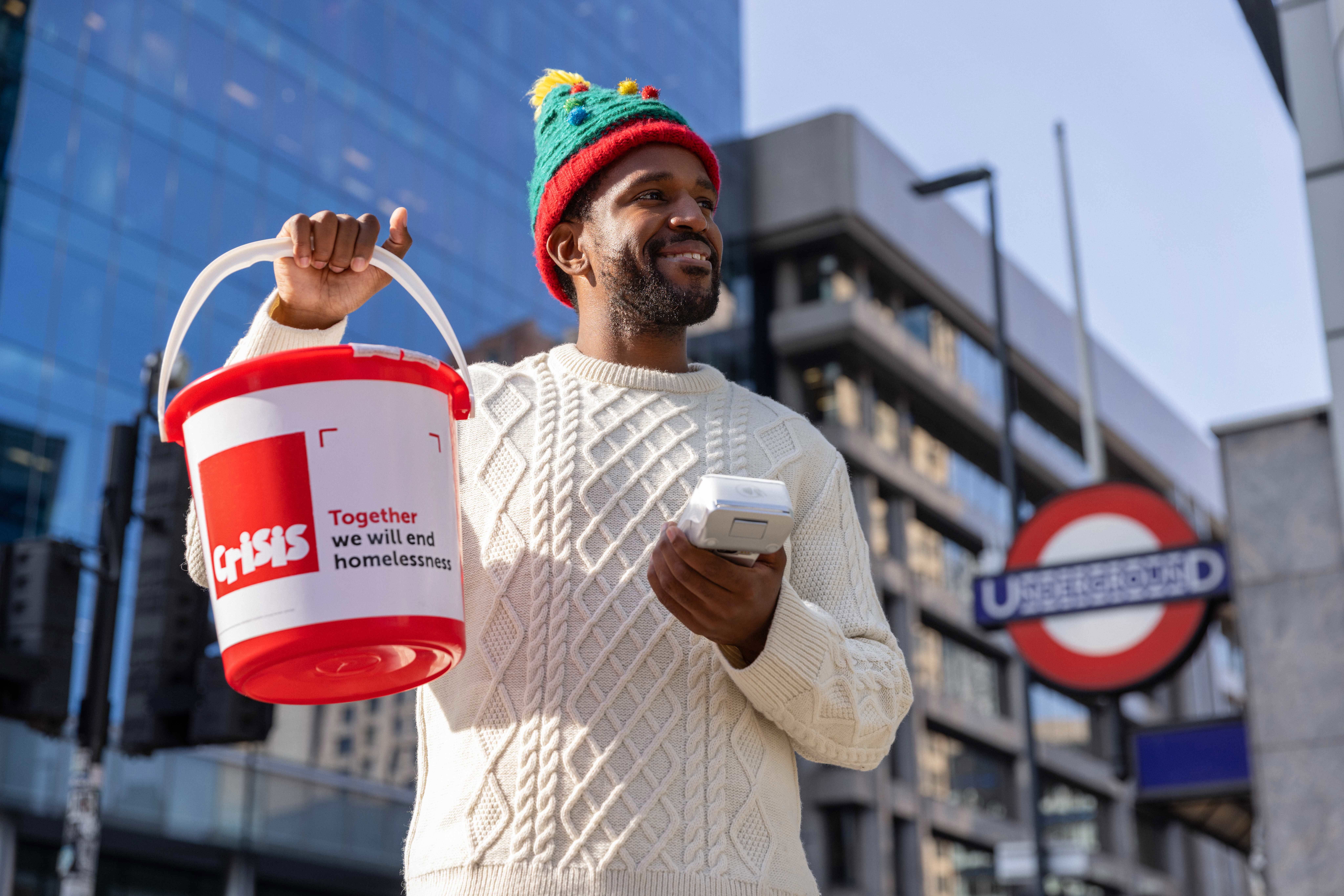 a man and woman standing in a street, holding a red Crisis collection bucket and smiling at the camera.
