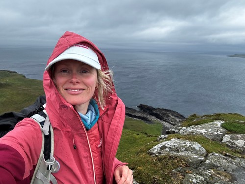A selfie of volunteer, Lucy. Lucy is standing on a coastal path, wearing a pink rain jacket and a white baseball cap. There is a sea view behind her.