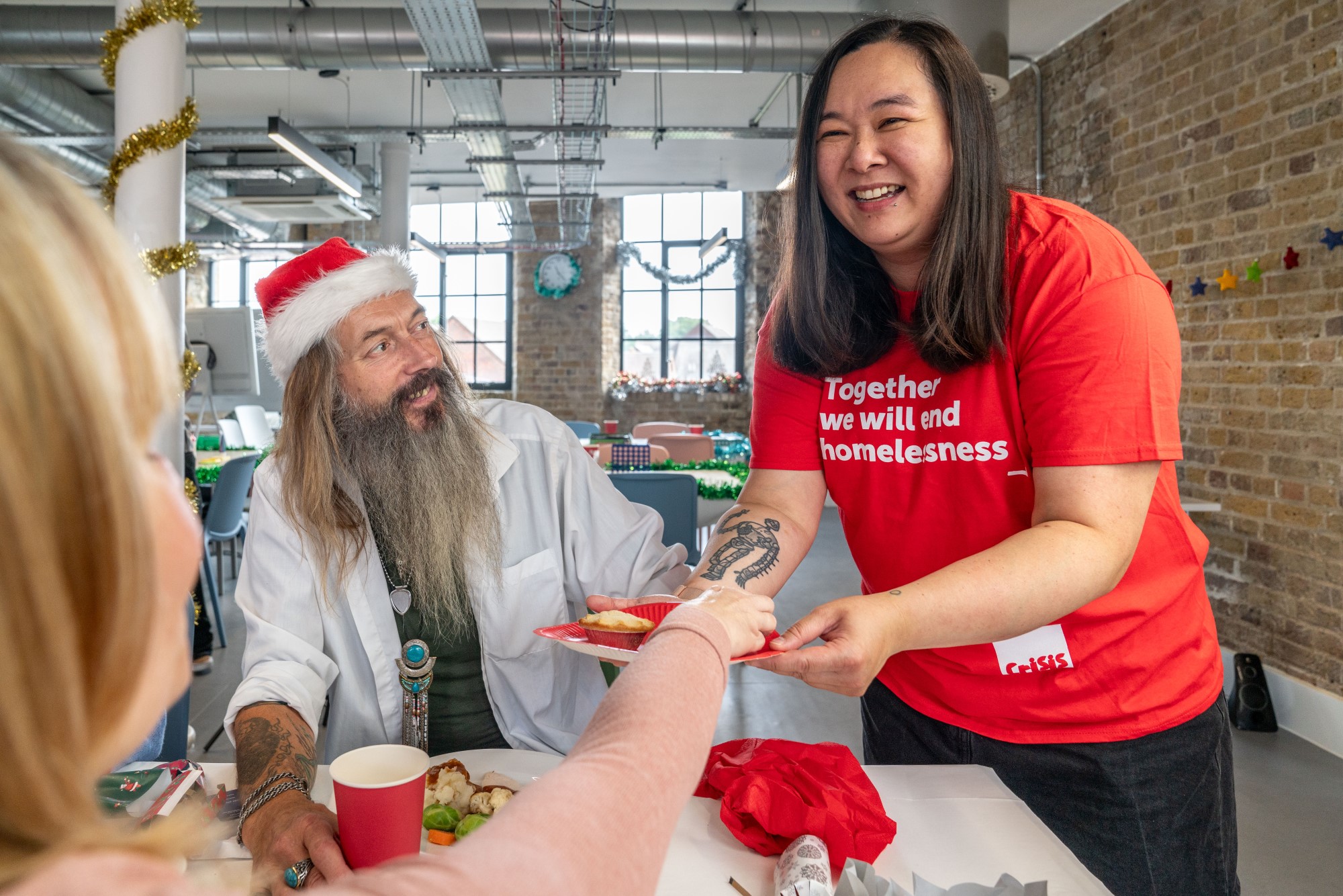 A woman wearing a Crisis t-shirt offers a plate of mince pies to seated Christmas guests. 