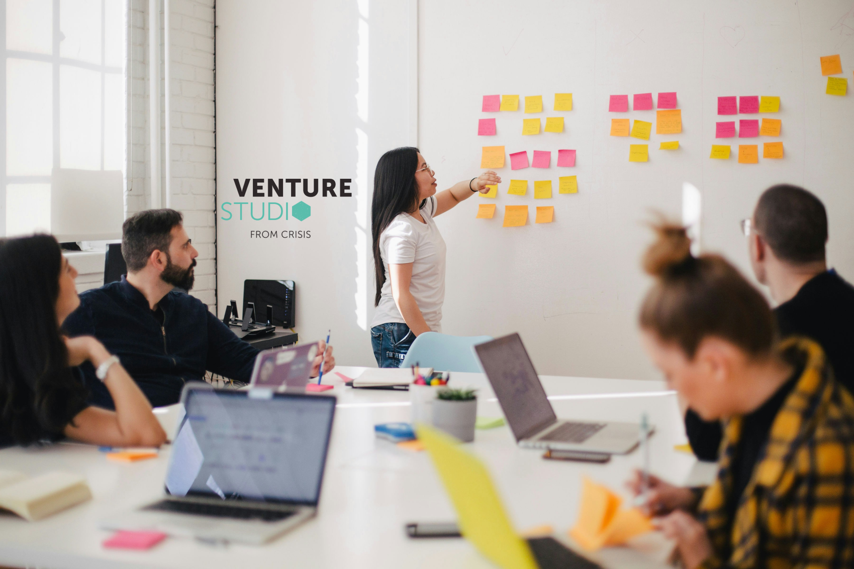 People sitting round a table in a meeting with one person presenting using post-it notes on a wall. 