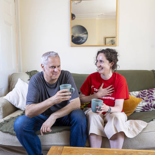 Massimo and a Crisis staff member are sat on a comfortable sofa. They are smiling and drinking tea. 